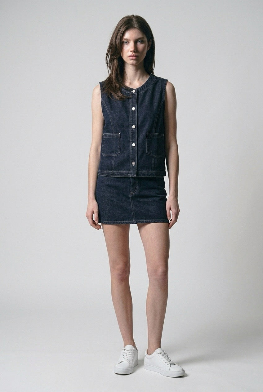 Front view of The Halo Denim Vest, collarless indigo denim with silver-tone buttons, styled with a denim mini skirt